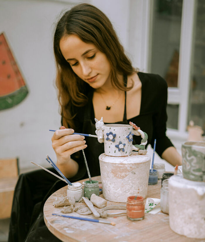 A young woman in a black apron meticulously painting a ceramic mug. The image implies expensive habits that save you money.