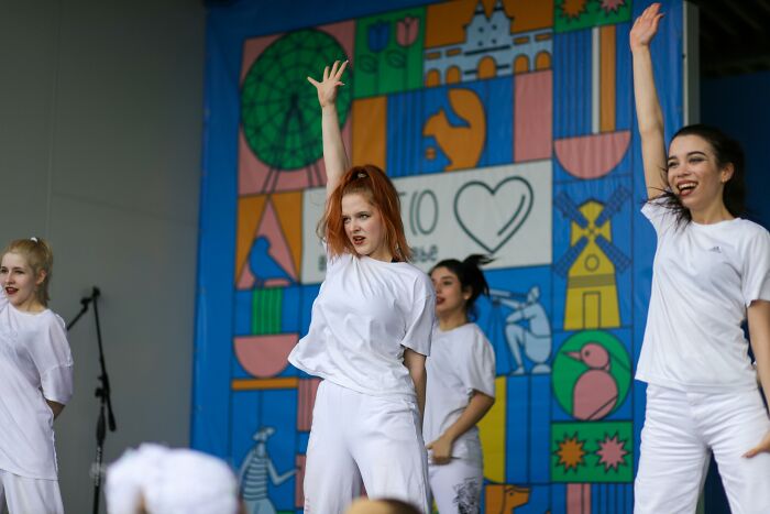 A group of young women in white outfits performing on stage, showing expressions of intense effort. Causes second-hand embarrassment.