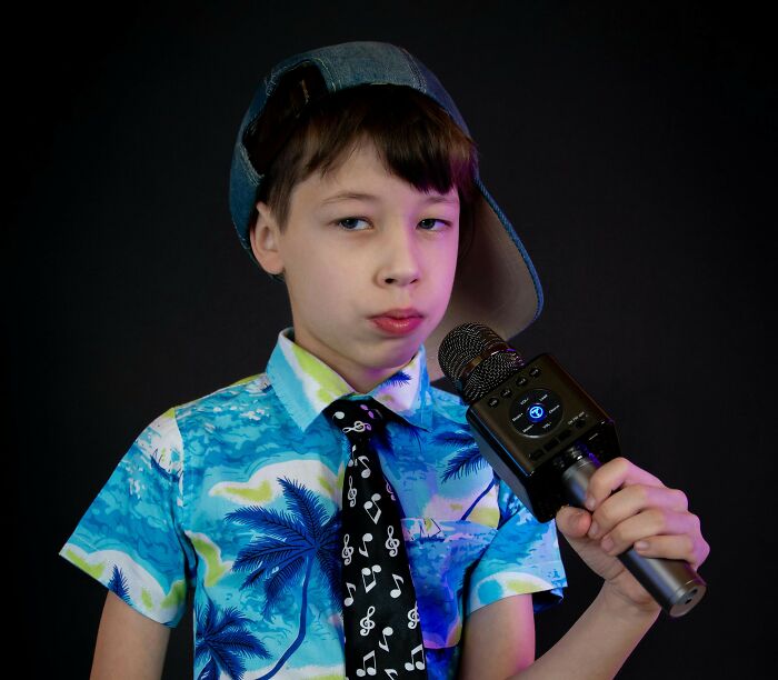 A young boy in a backward cap, Hawaiian shirt, and music note tie holds a karaoke microphone, looking slightly cringe.