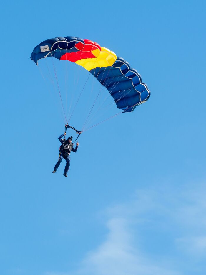 A skydiver descends under a blue, red, and yellow parachute against a clear blue sky, experiencing something creepy.