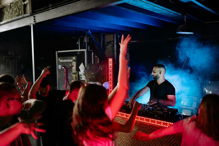 A DJ performing at a lively party with Gen Z dancing in front of the stage, blue smoke and red lights.