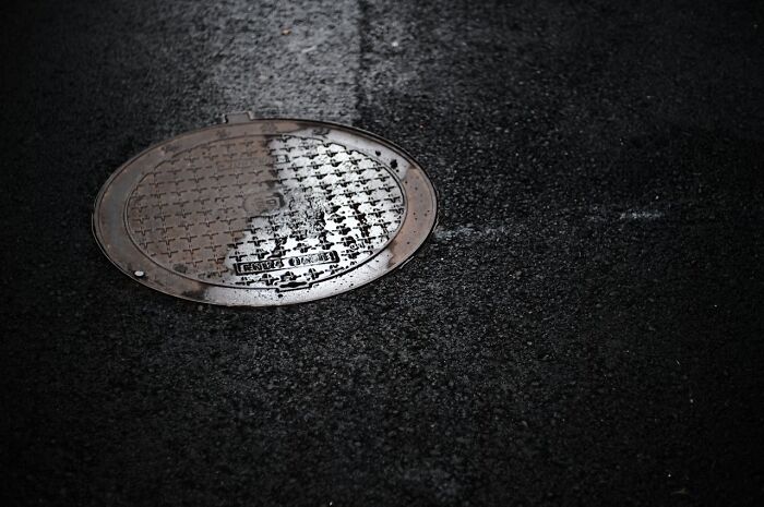 A wet manhole cover on dark asphalt, reflecting light; a subtle detail in d***h stories imagery.