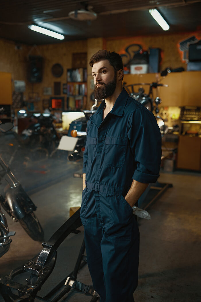 Bearded mechanic standing in workshop surrounded by tools and motorcycles