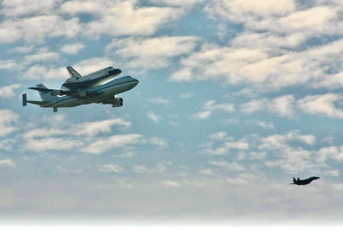 Space shuttle mounted on airplane flying alongside fighter jet under cloudy sky