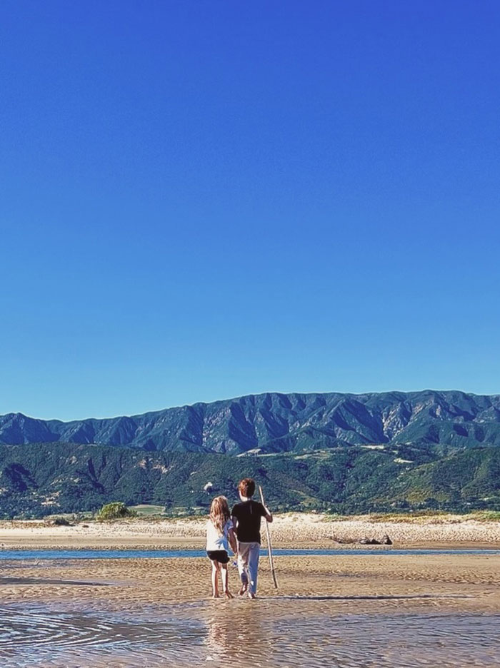 Children on a beach, facing distant mountains under a clear sky. A boy holds a stick. Meghan Markle and Prince Archie.