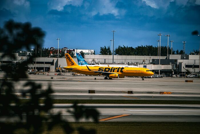 A bright yellow Spirit Airlines plane on a tarmac, with a JetBlue plane behind it, at an airport under a blue sky.