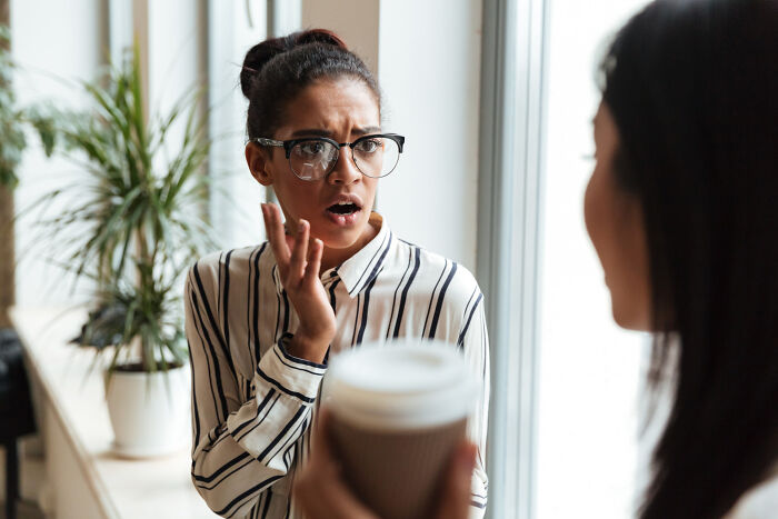 A woman with glasses and a striped shirt looks surprised, revealing a tiny red flag. Another person offers coffee.