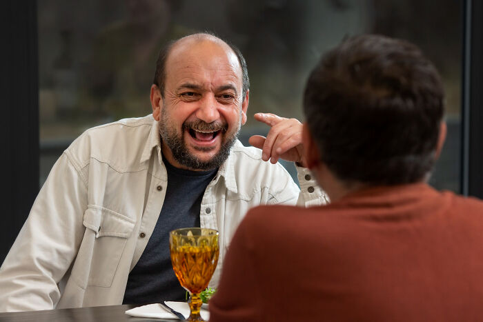 Two men at a table, one in white shirt laughing heartily, the other seen from behind. Reveals more about someone.
