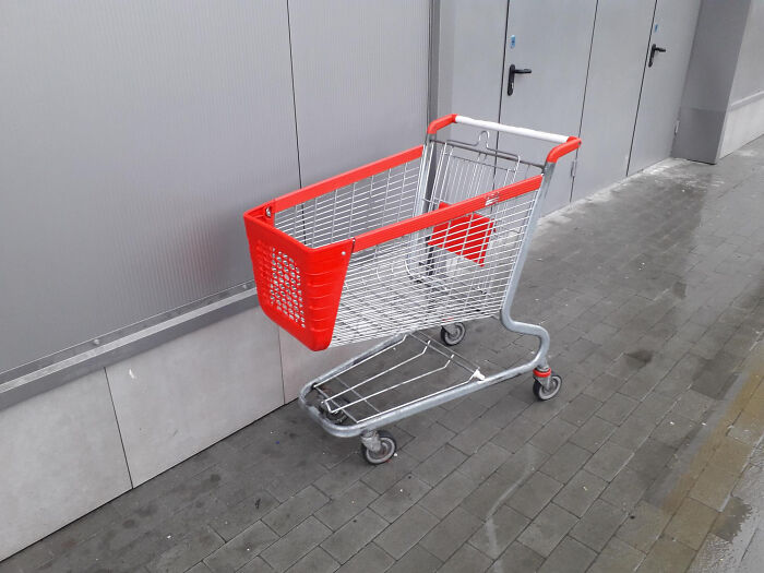 A shopping cart with a bright red basket and silver frame sits abandoned on a wet grey sidewalk. This could be a tiny red flag.