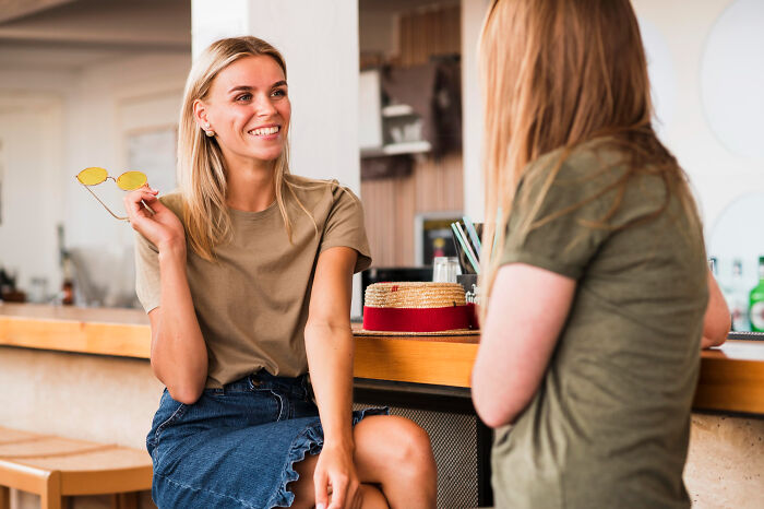 Two women chatting at a bar. One with blonde hair smiles, holding yellow sunglasses. Tiny red flags are discussed.
