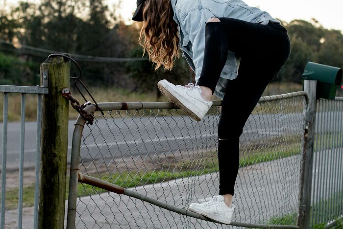 A young woman in a denim jacket and ripped jeans climbs a chain link fence, revealing red flags of risky behavior.