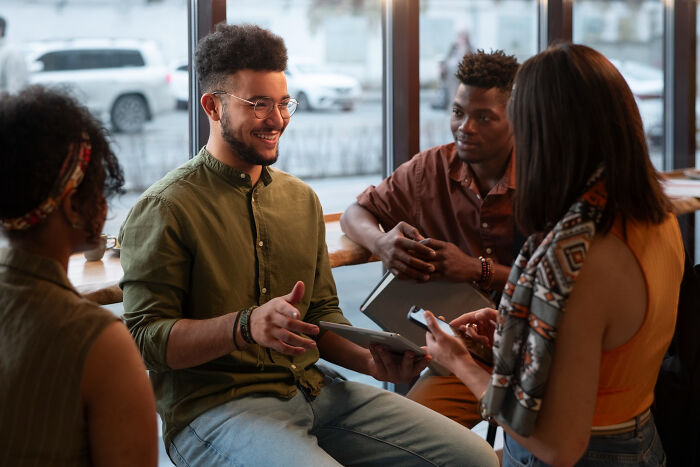 Four diverse people in a cafe, a man holding a tablet and smiling. Discussing tiny red flags about someone.