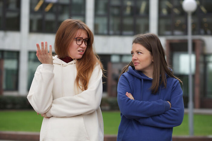 Two young women, one gesturing with a confused expression, the other listening intently. A subtle tiny red flag in their interaction.