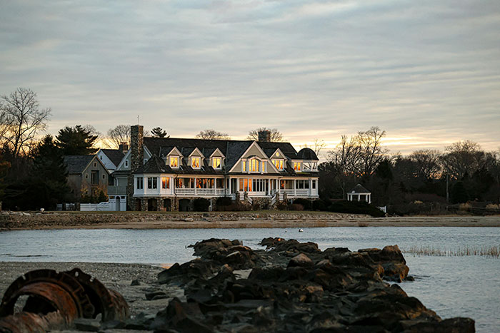 A large, luxurious house with many windows lit up at dusk, situated on a waterfront. Represents the ultra-rich lifestyle.