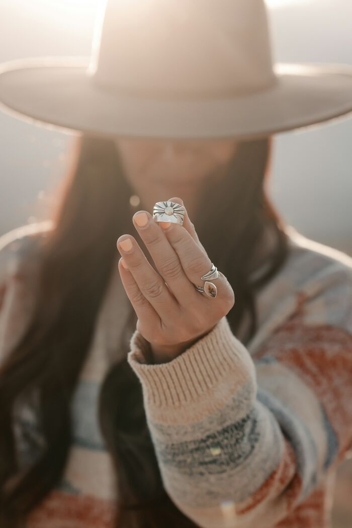 Woman wearing a hat and patterned sweater, holding out a hand with rings, one a silver sun, questioning reality.