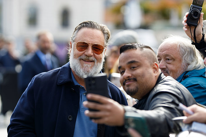 Russell Crowe smiling and taking selfie with fan in friendly crowd scene