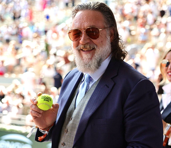 Russell Crowe smiling in sunglasses holding a tennis ball at a public event