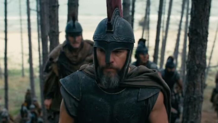 Homem barbudo com armadura e capacete romanos, com outros soldados, em uma floresta, para A Odisséia, de Christopher Nolan.