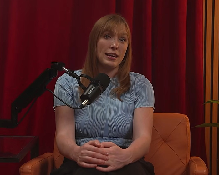 A woman with reddish-brown hair and a light blue top sitting in a chair, talking into a microphone, discussing Sabrina Carpenter Catfish accusations.
