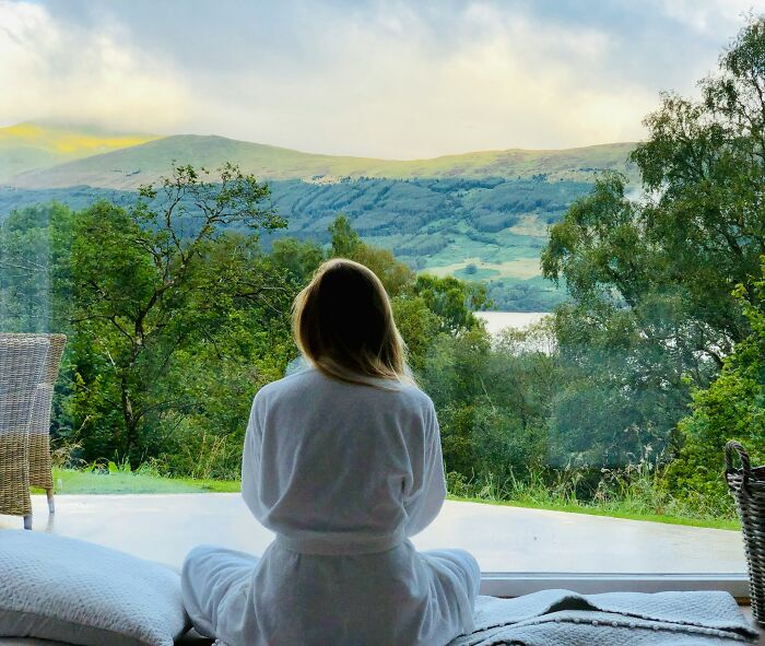 Woman in a robe enjoying silent luxuries, meditating by a window with a serene mountain and lake view.