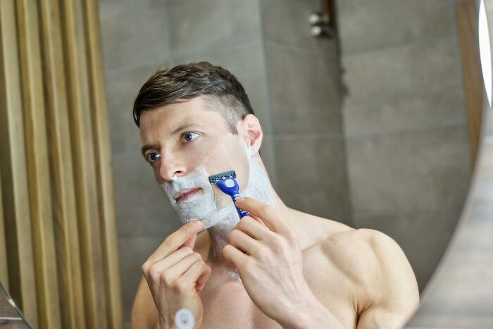 A man with shaving cream on his face, looking in a mirror while shaving. Many outdated facts about shaving exist.