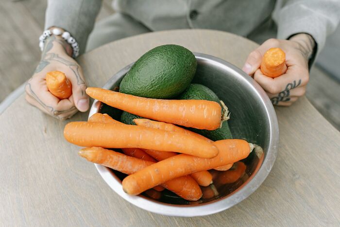 A person with tattooed hands holding carrot sticks, with a bowl of carrots and avocados. Challenging outdated facts about diet.