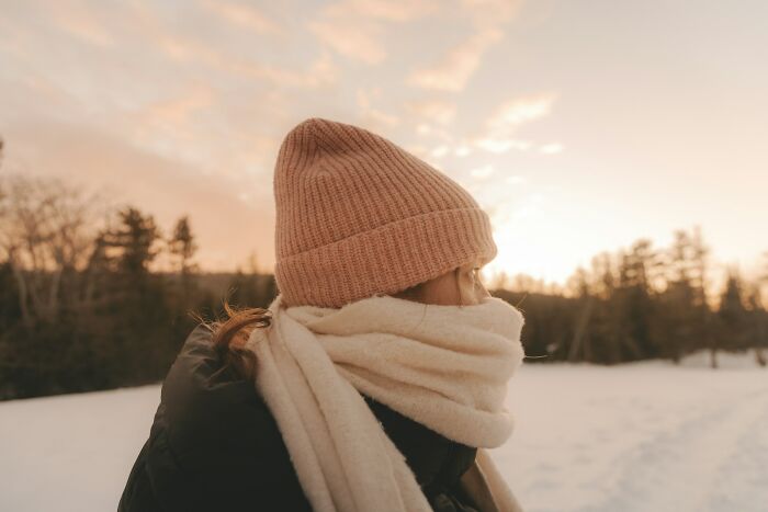 A person in a pink beanie and white scarf looks out at a snowy, tree-lined landscape under a sunset sky, contemplating outdated facts.