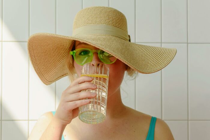 Woman in a straw hat and green sunglasses drinking water, challenging outdated facts about hydration and health.