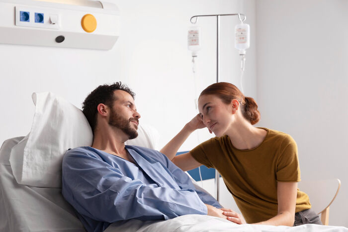 A woman leaning on a hospital bed, looking at a man in a patient gown. Both gaze at each other. Keeping secrets from significant others.