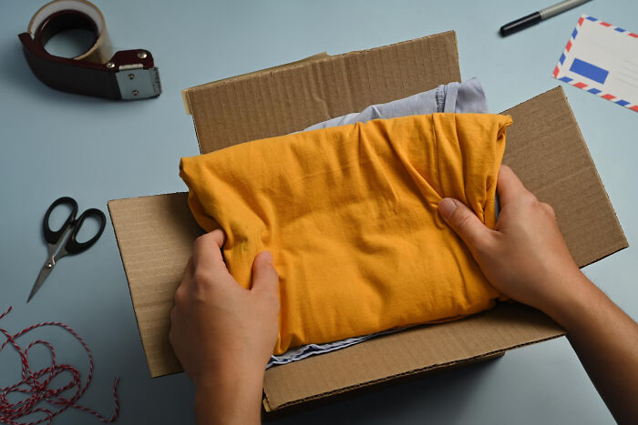 Hands placing a folded orange shirt into a cardboard box. This image may relate to keeping secrets or sending a package.