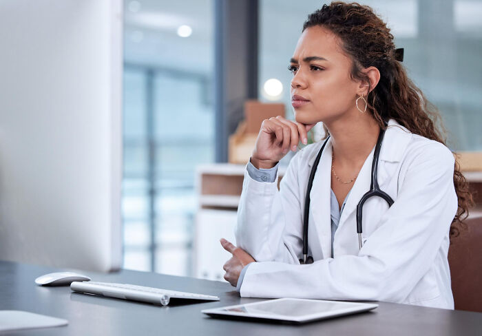 A thoughtful female doctor in a white coat and stethoscope, looking at a computer screen. Keeping secrets, honest confessions.