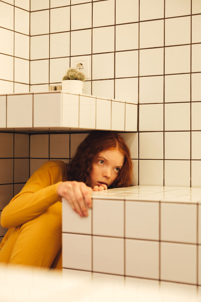 A young woman with red hair looking shocked, hiding behind a tiled surface. A metaphor for someone finding out their partner was cheating.