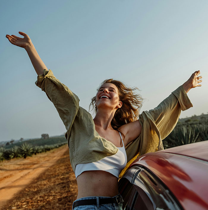 Joyful woman with raised arms near car enjoying outdoors beauty lies uncovered