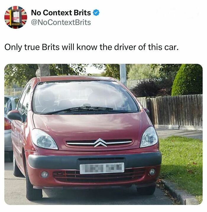 A red Citroen car, featured in a British meme, parked on the street with trees and a wooden fence in the background.