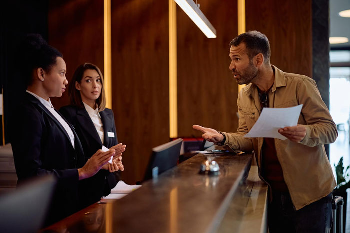 Man confidently speaking to two female employees at front desk in workplace setting Man confidently speaking to two female employees at front desk in workplace setting