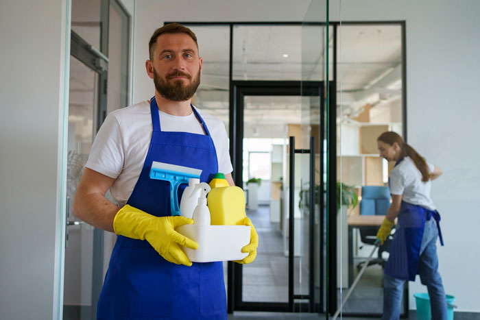 Male cleaner, wearing a blue apron and yellow gloves, holds cleaning supplies. Another cleaner mops in the background. Family driver.