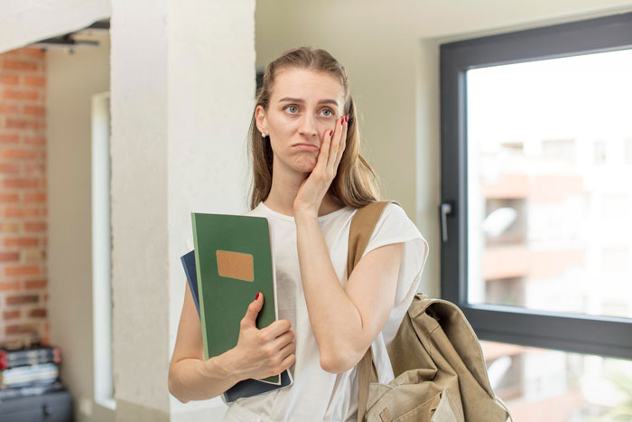 A young woman, a 20YO, looking sad and embarrassed, holding books and wearing a backpack, contemplating losing her funds.