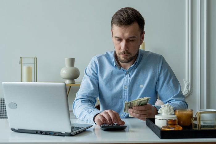A man in a blue shirt counting money and using a calculator at a desk with a laptop, reflecting the keyword 'funds'.