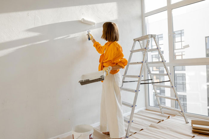 A person in an orange shirt and white pants redecorates a room by painting a white wall with a roller. Sister surprise redecoration.