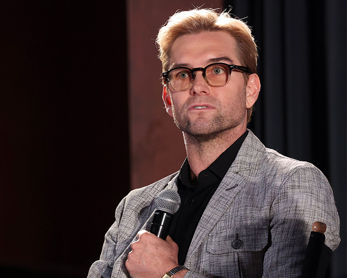 The Boys star speaking at event holding microphone with glasses and blazer The Boys star speaking at event holding microphone with glasses and blazer