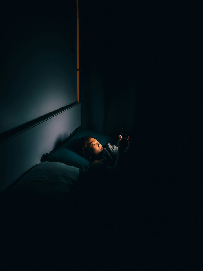 A woman in bed, face lit by a phone screen in a dark room. She looks at her device, likely smart scale related.