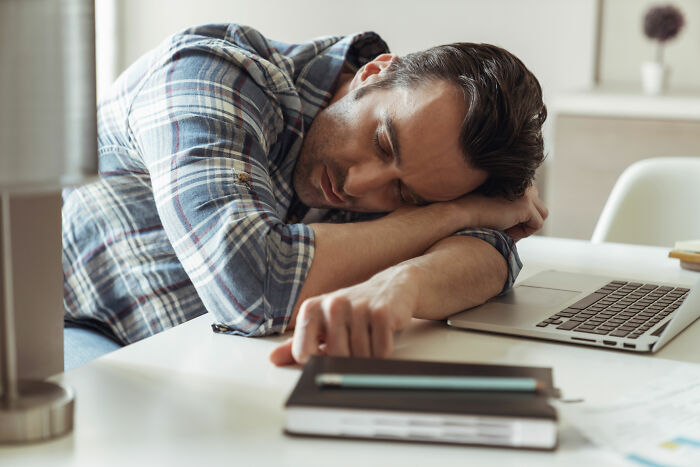 A tired man in a plaid shirt rests his head on his arms at a desk with a laptop, highlighting fatigue as a sign for routine cancer screening.