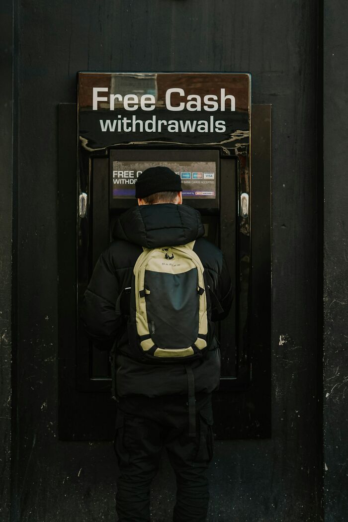 A person in a black jacket and backpack uses an ATM displaying Free Cash withdrawals. Visiting the US moments.