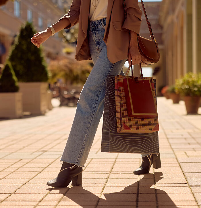 A person in jeans and a blazer carrying shopping bags on a sunny street, exploring habits that save money.