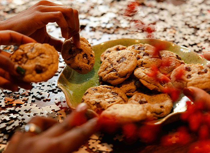 Hands reaching for chocolate chip cookies from a green plate, with jigsaw puzzle pieces scattered. Unwritten rules, Brits.
