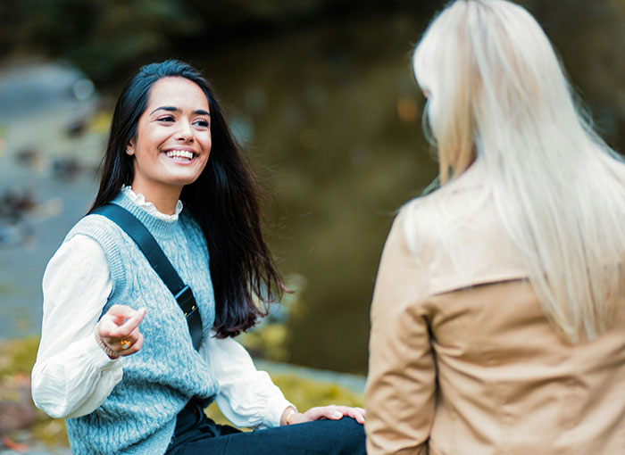 A smiling young woman with long dark hair, wearing a white shirt and grey vest, talking with another person. Brits unwritten rules discussion.