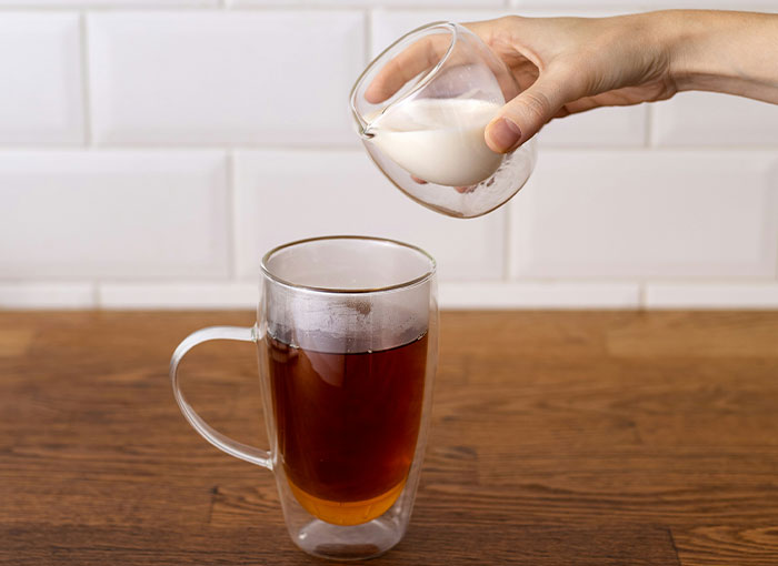 A hand pours milk into a glass mug of tea, reflecting British tea-drinking rules and traditions.