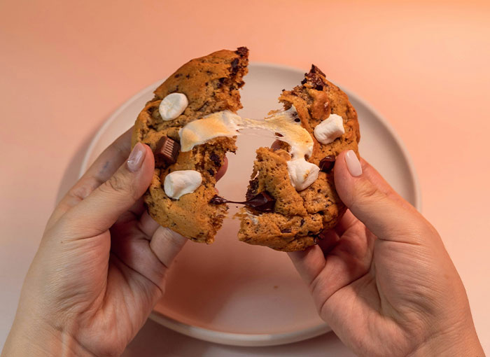 Hands pulling apart a warm chocolate chip and marshmallow cookie, with melted marshmallow stretching between the two halves. #Brits #UnwrittenRules