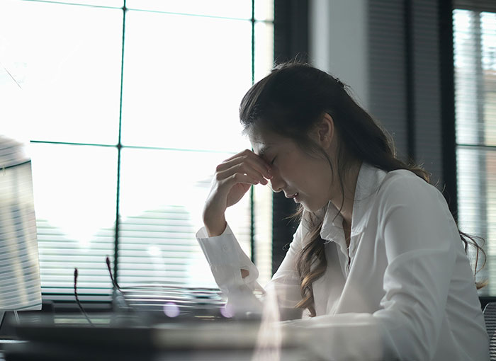A stressed woman, hand on her forehead, at a desk by a window. Explores unwritten rules Brits share.