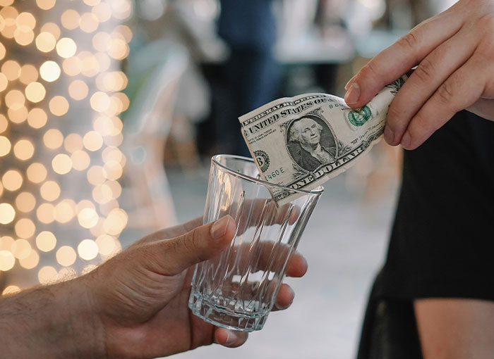 A person puts a dollar bill into an empty glass held by another, highlighting unwritten rules, maybe tipping in Britain.